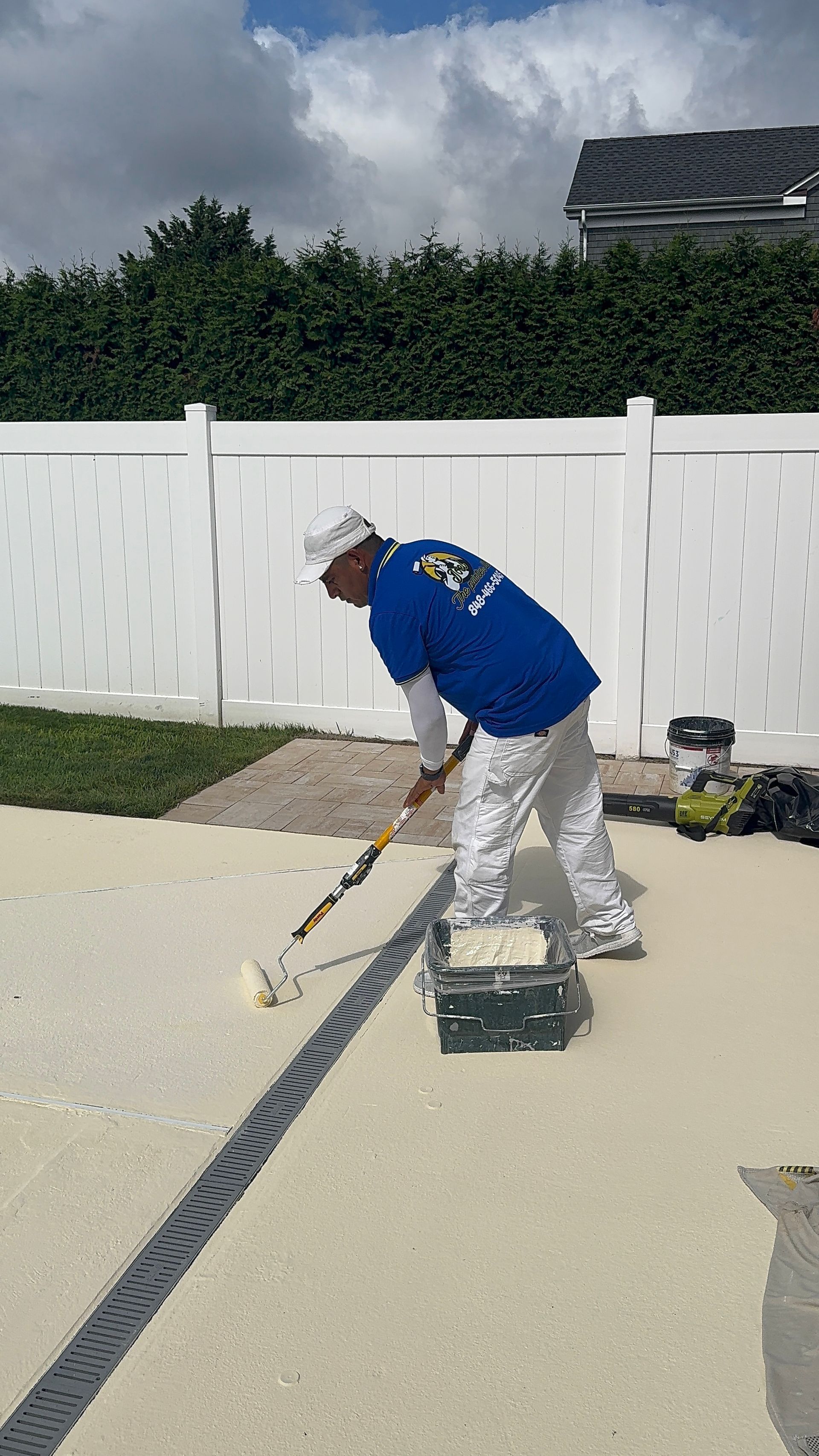 Worker painting a concrete driveway with a roller near a white fence on a sunny day