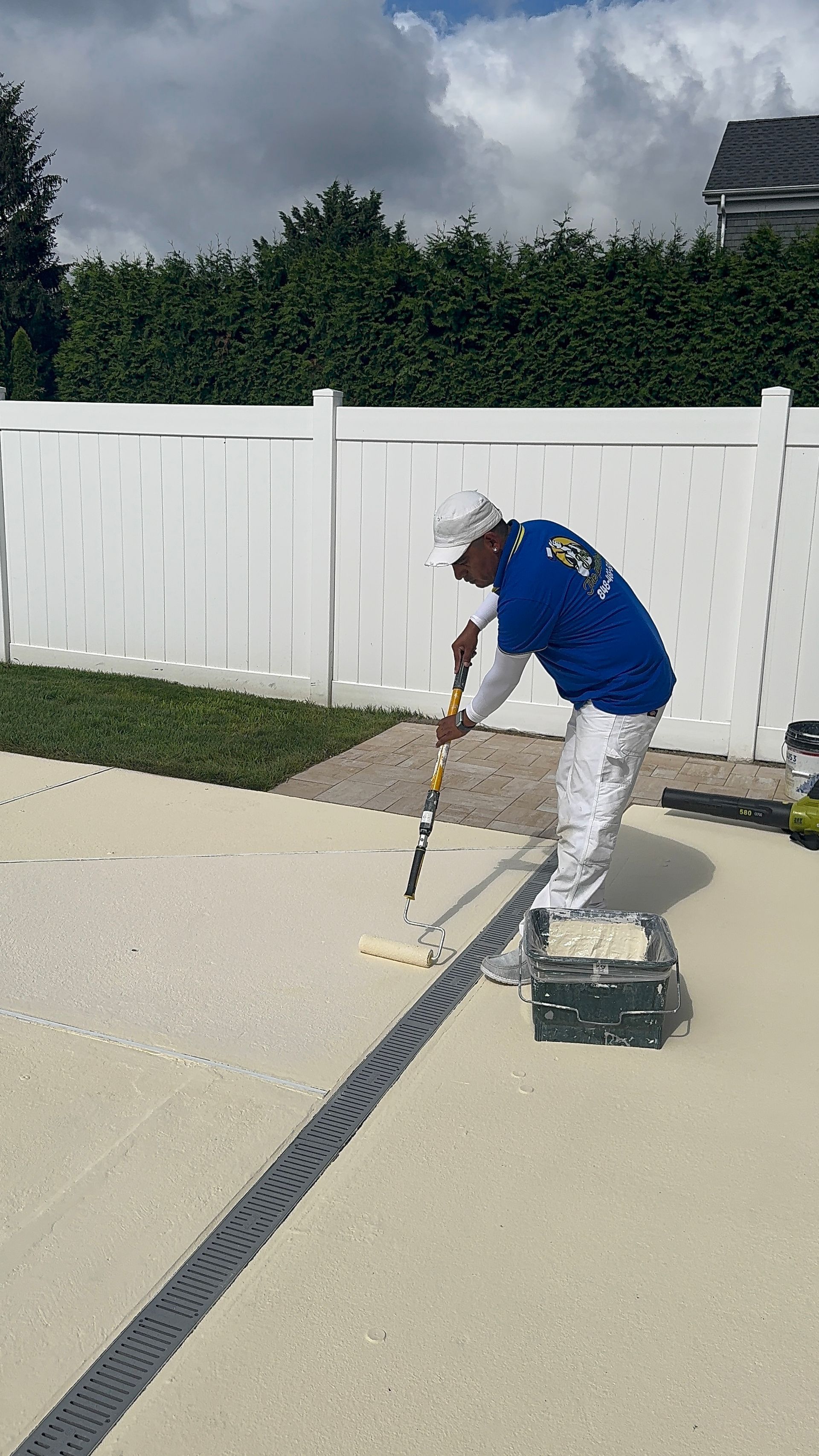 Worker painting a driveway with a roller beside a white fence and hedge.