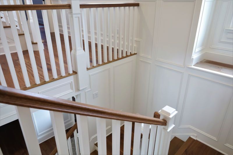 Bright stairwell with white paneled walls, wooden handrails, and white balusters by a window.