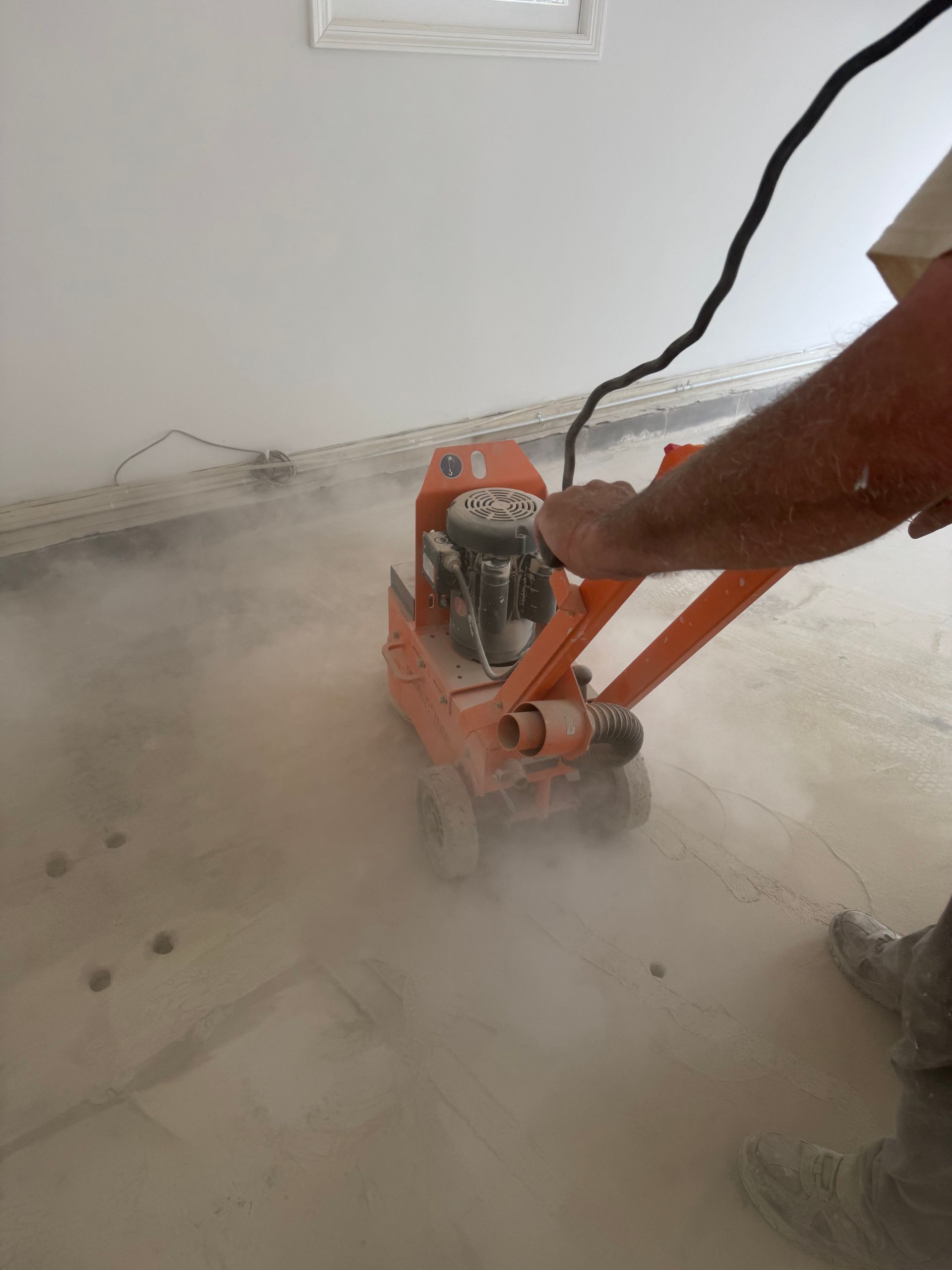 Worker using an orange floor grinder, creating heavy dust in an indoor room with bare walls.
