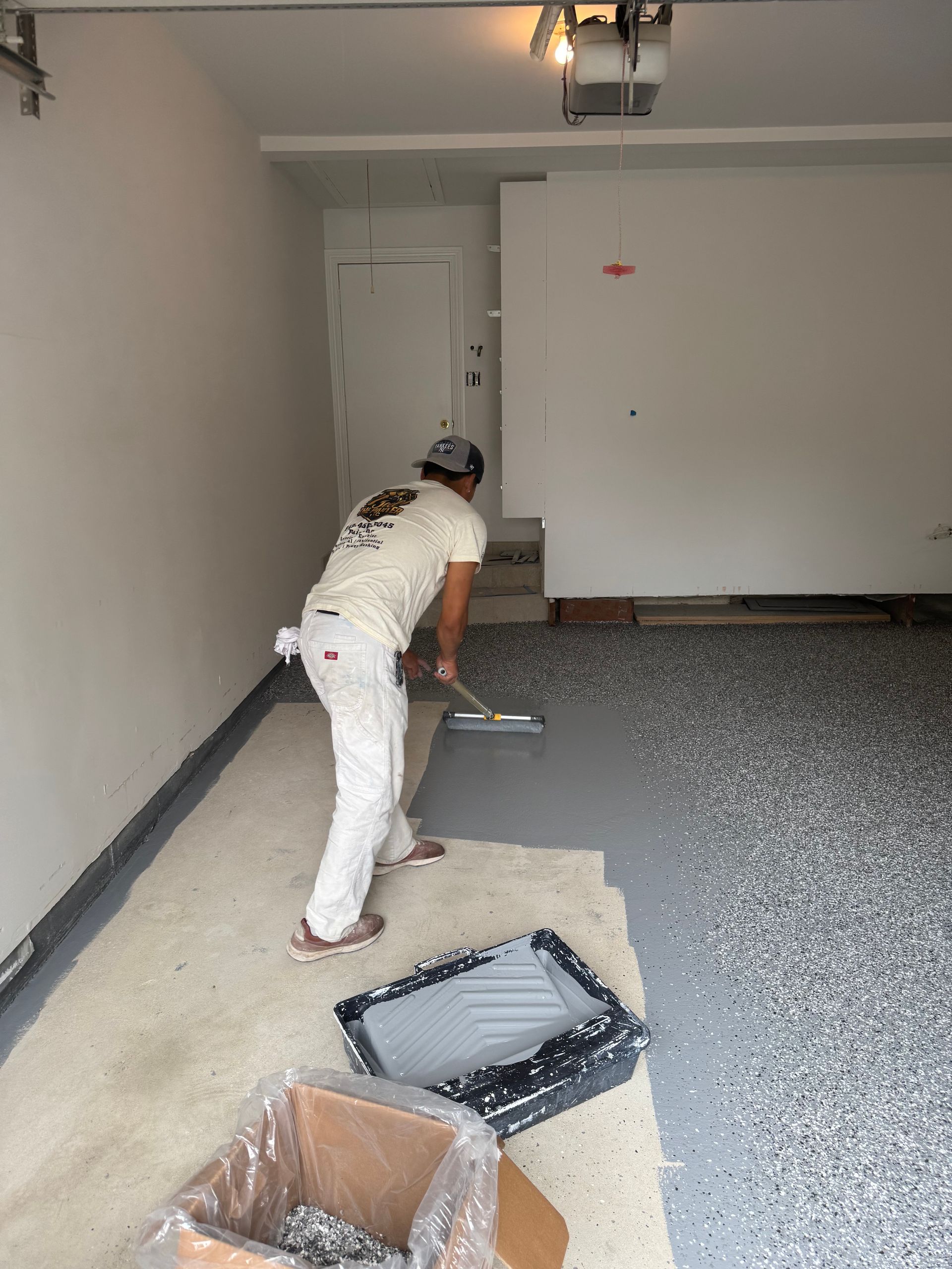 Person painting a garage floor, with paint trays and supplies on the ground.