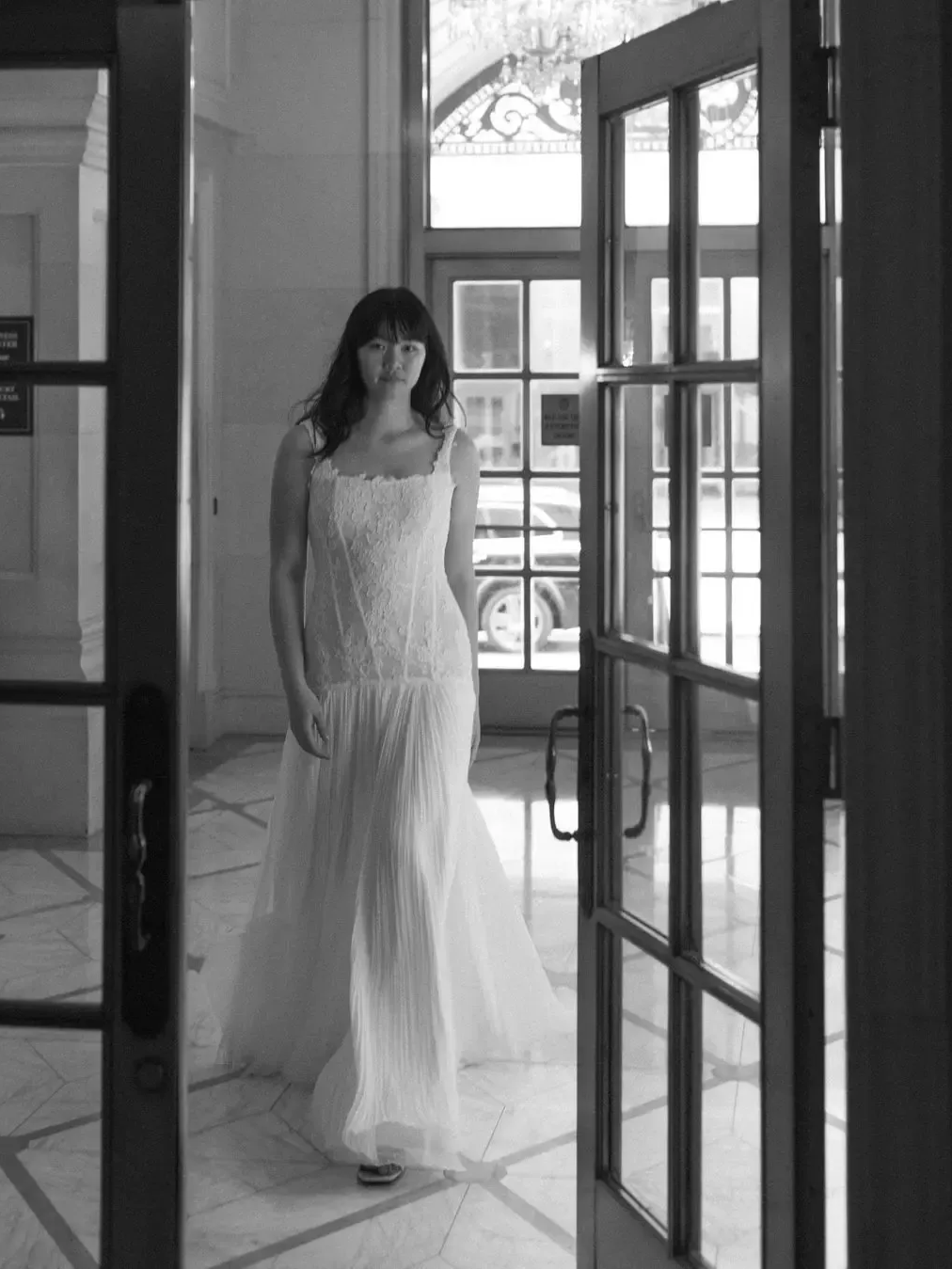 Woman in wedding dress, standing in a doorway. Interior hallway with marble flooring and large windows. Black and white.
