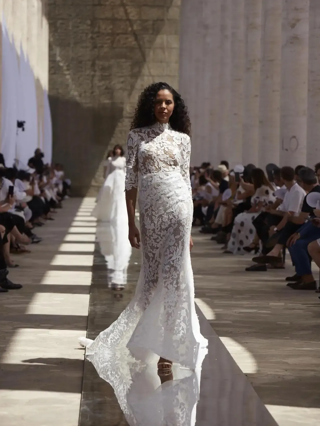 Model in a white lace wedding dress walks down a runway in an outdoor setting.