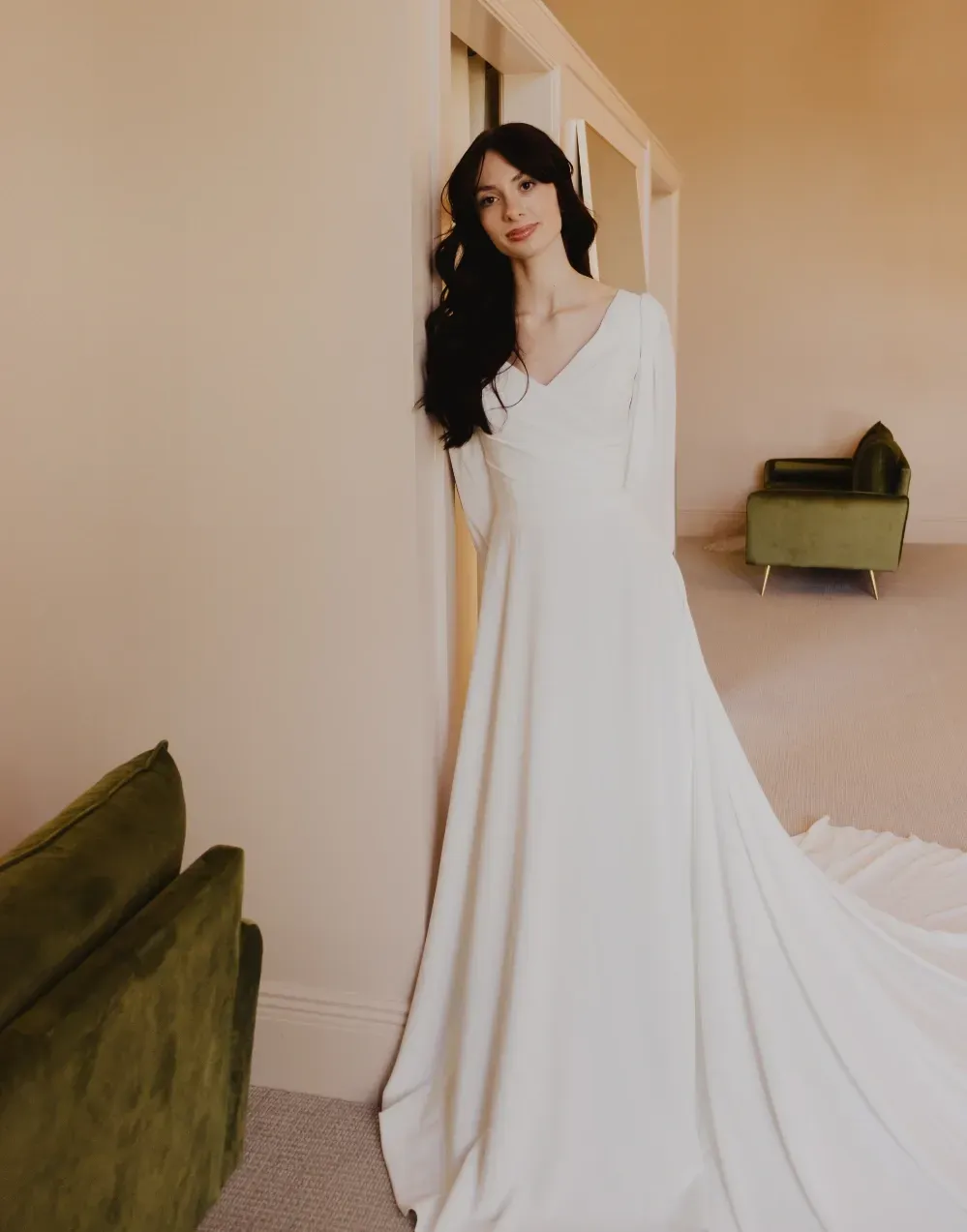 Woman in a white wedding dress leans against a wall in a room, long train, green chairs.
