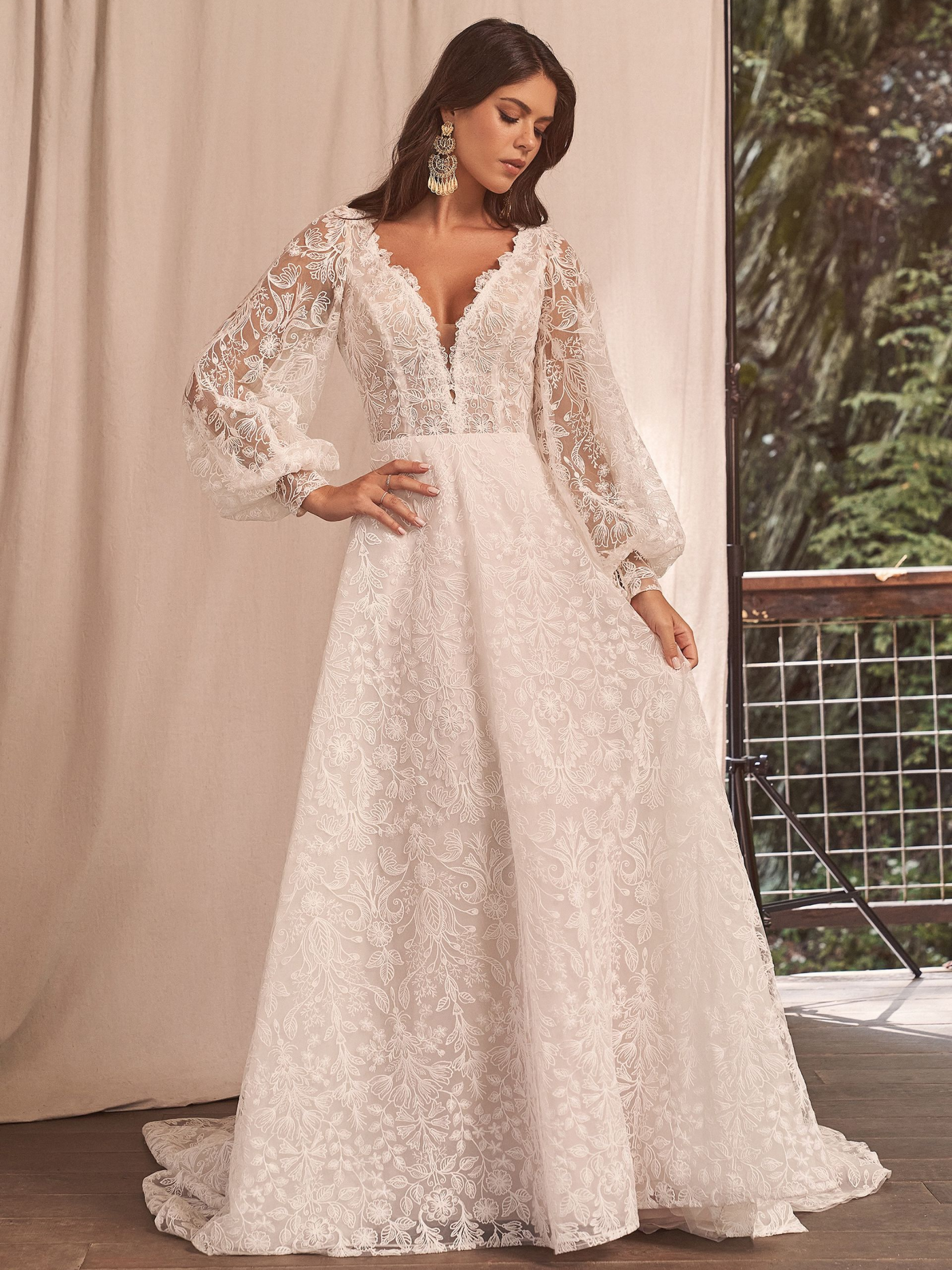 Woman in white, floral-embroidered wedding dress with long sleeves, posing outdoors.