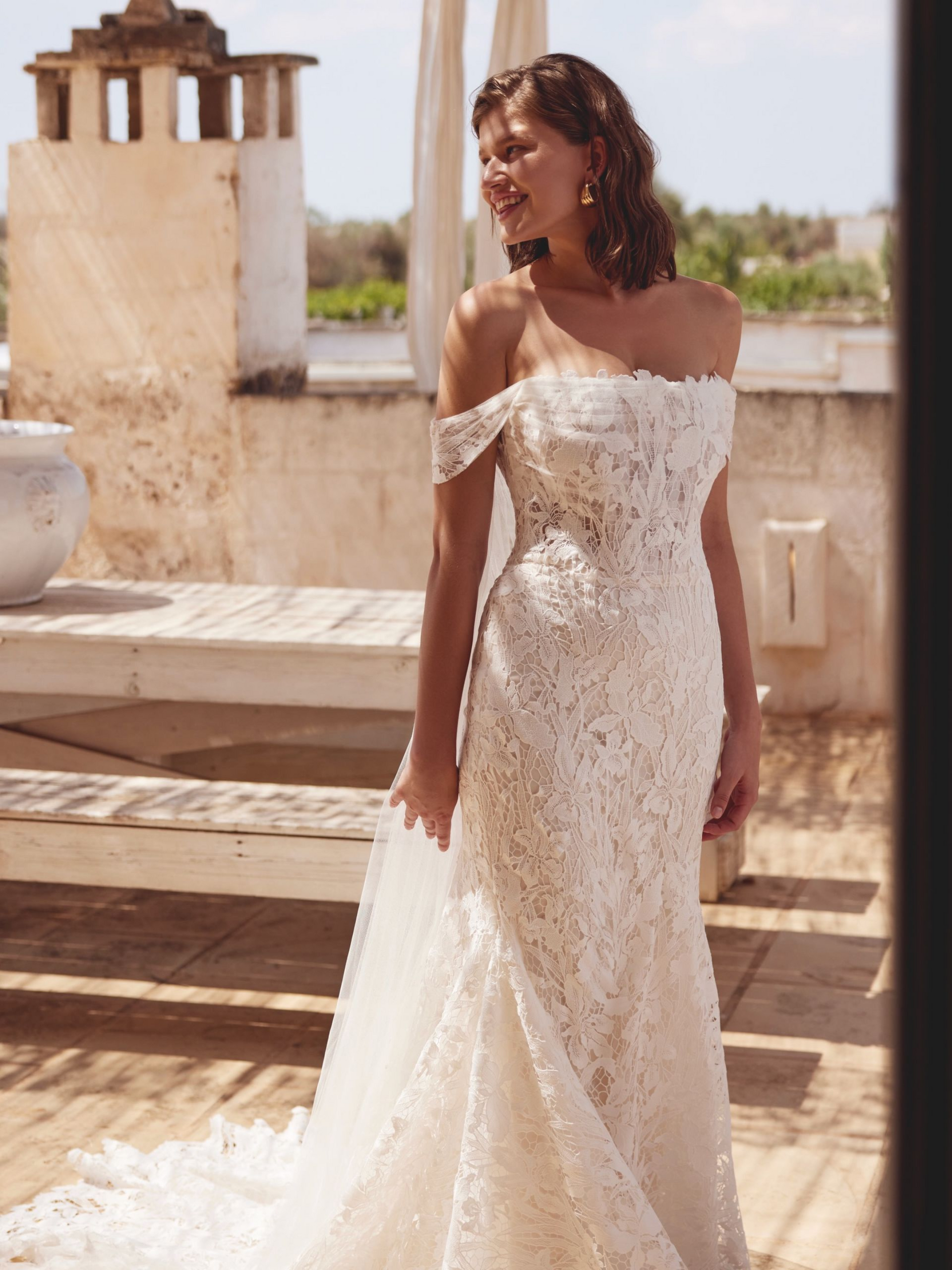 Woman in a lace off-the-shoulder wedding dress on a sunny terrace.