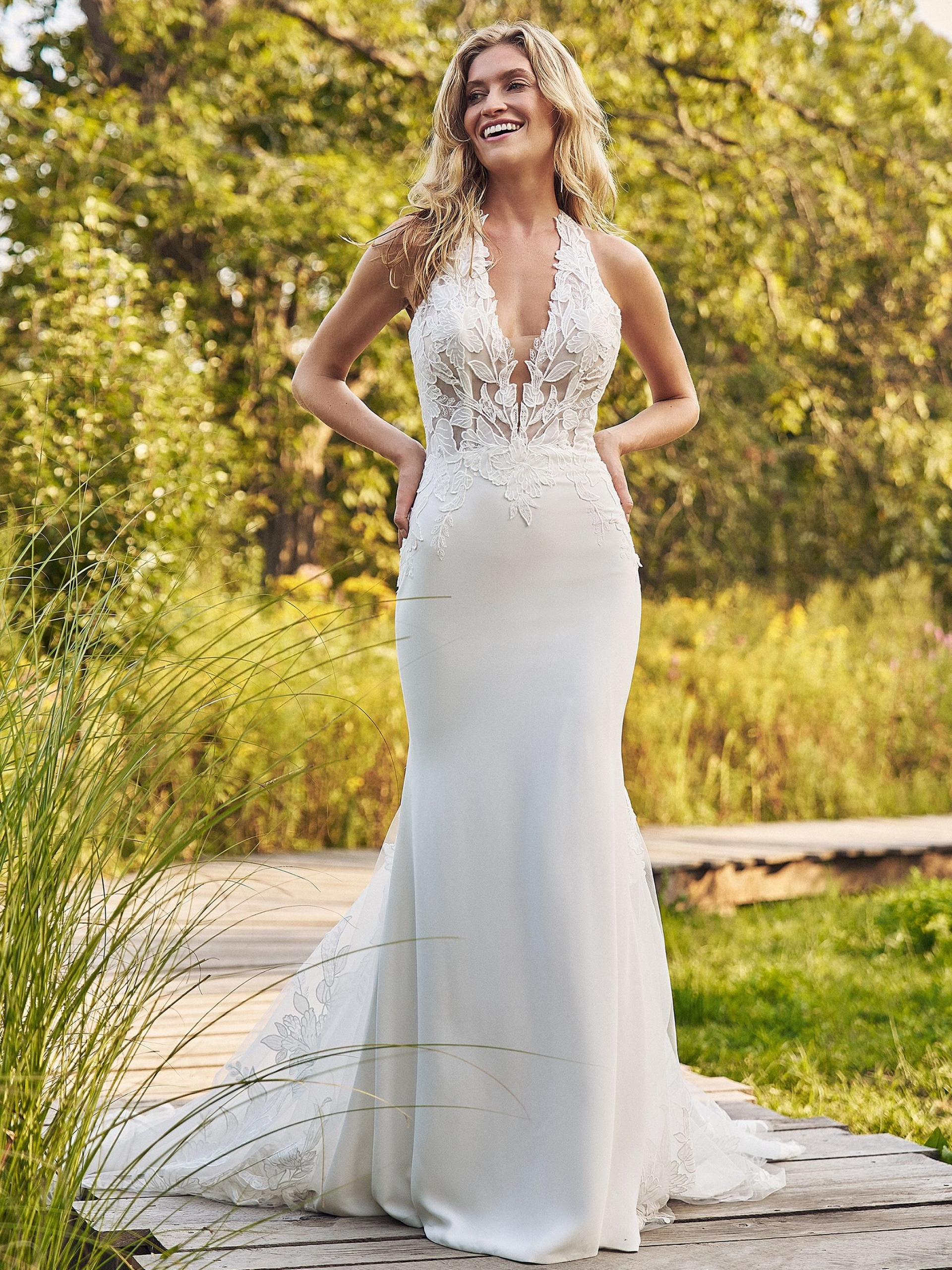 Woman in a white wedding dress with a deep v-neck, smiling outdoors, on a wooden bridge, green background.