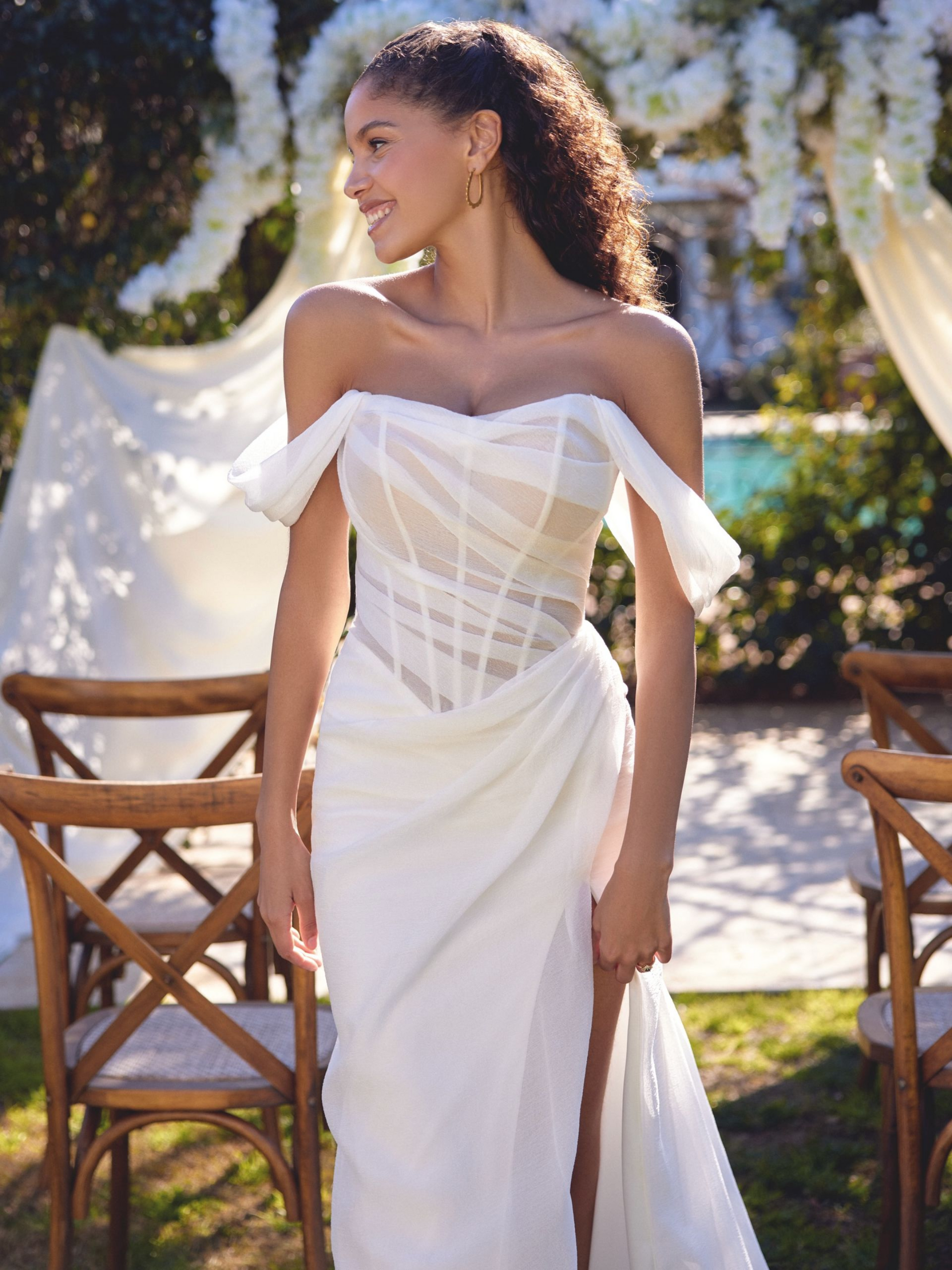 Woman in off-the-shoulder white wedding dress, smiling outdoors near ceremony setup.
