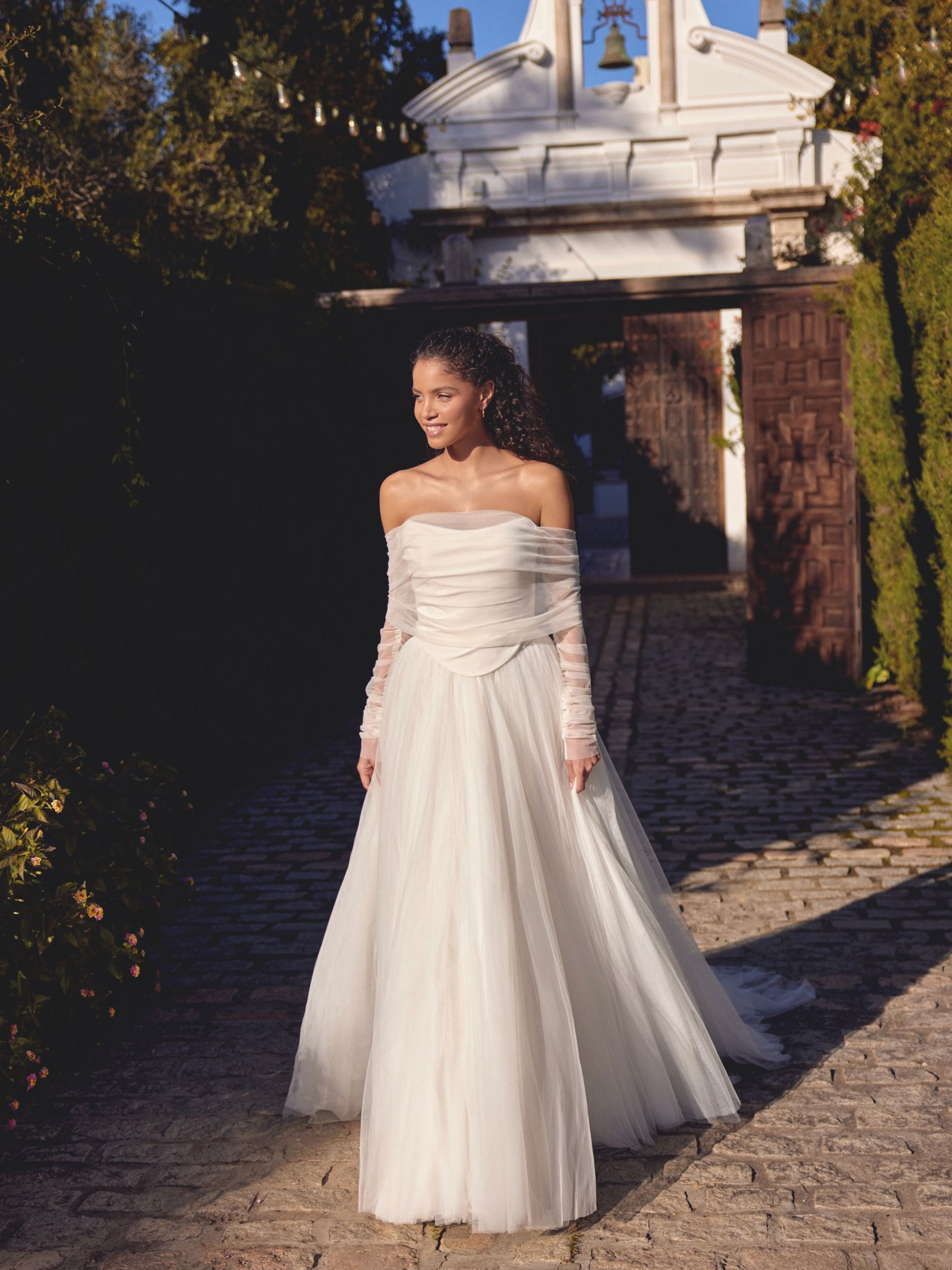 Woman in a wedding dress smiles, standing outdoors by a white ornate gate and a brick path.