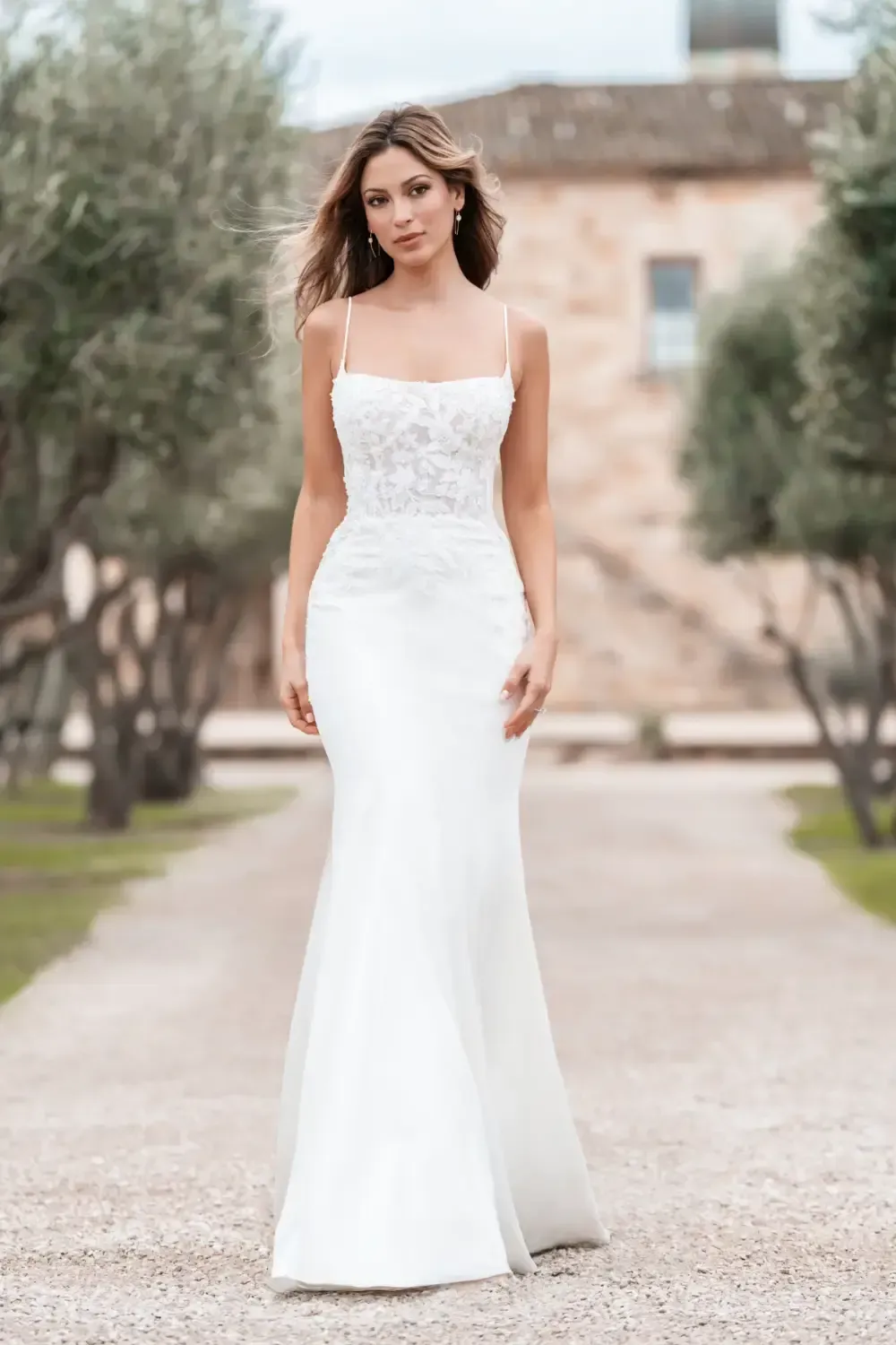 Woman in a white wedding dress stands on a stone path, with trees and a building in the background.
