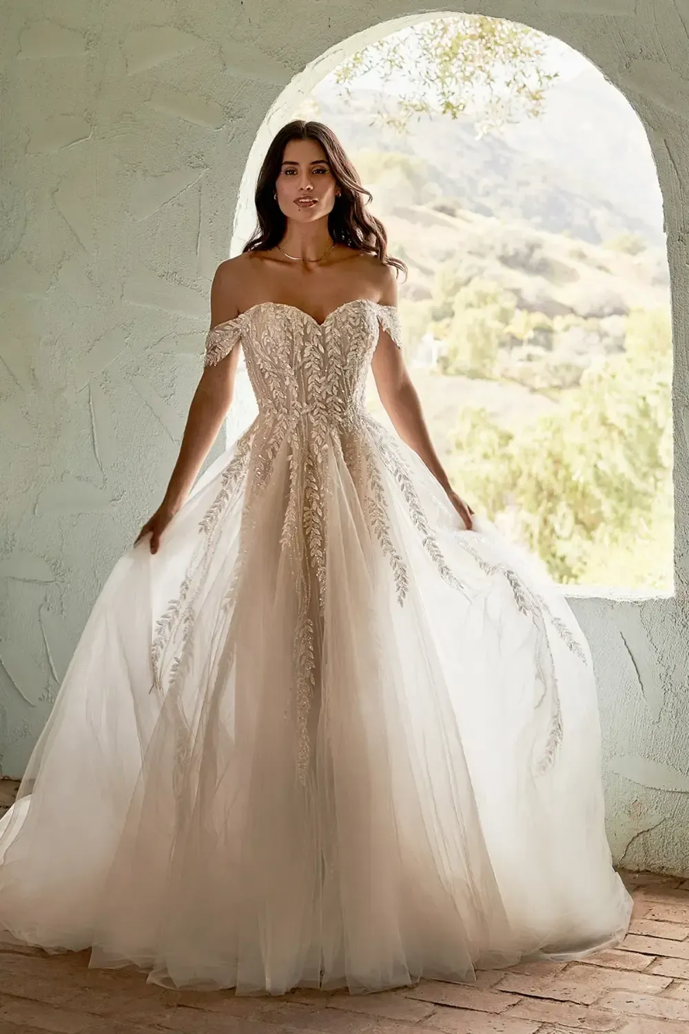 Woman in off-the-shoulder wedding dress, standing near an arched window, holding out skirt.