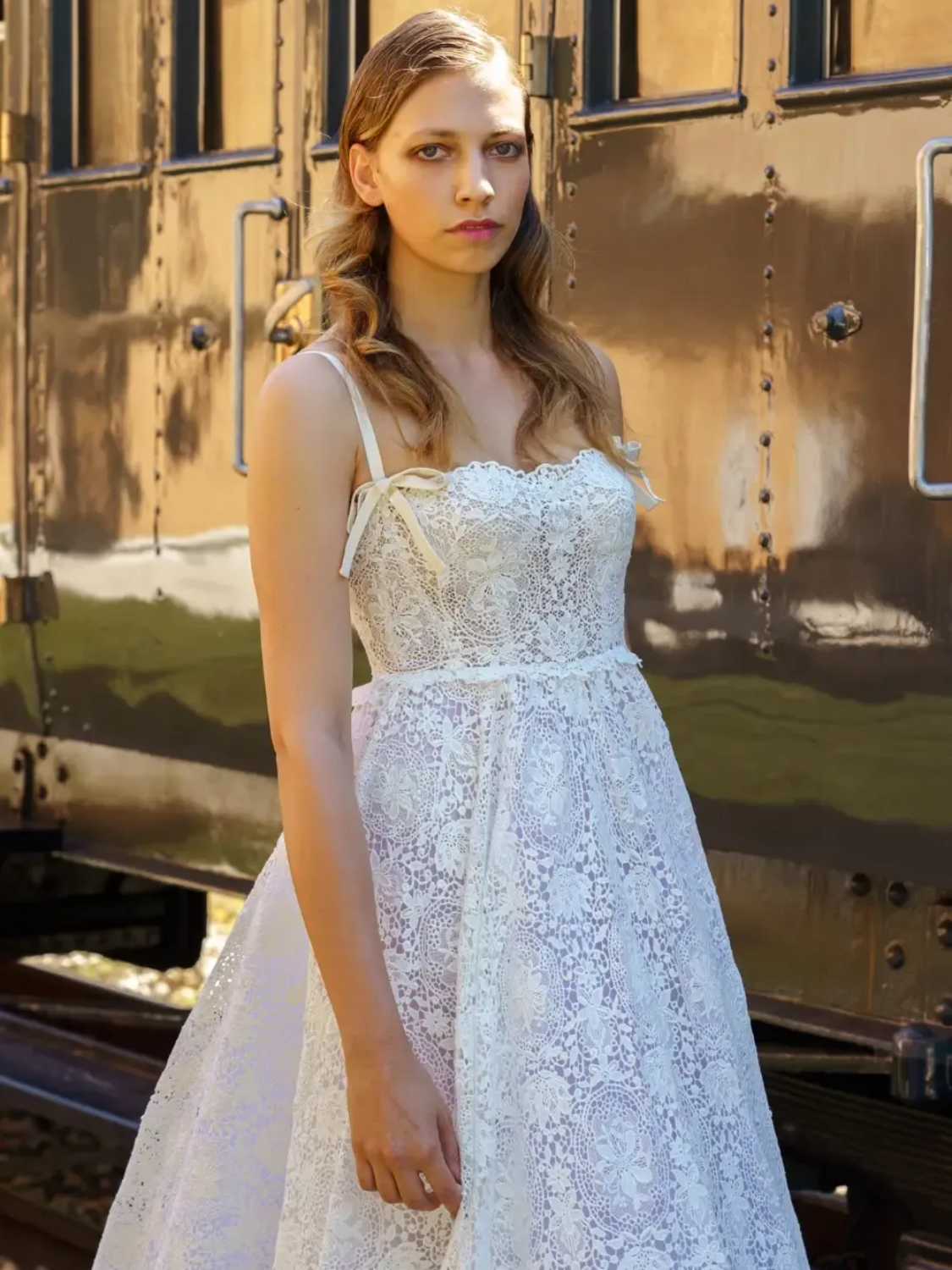 Woman in white lace dress poses by a train car.