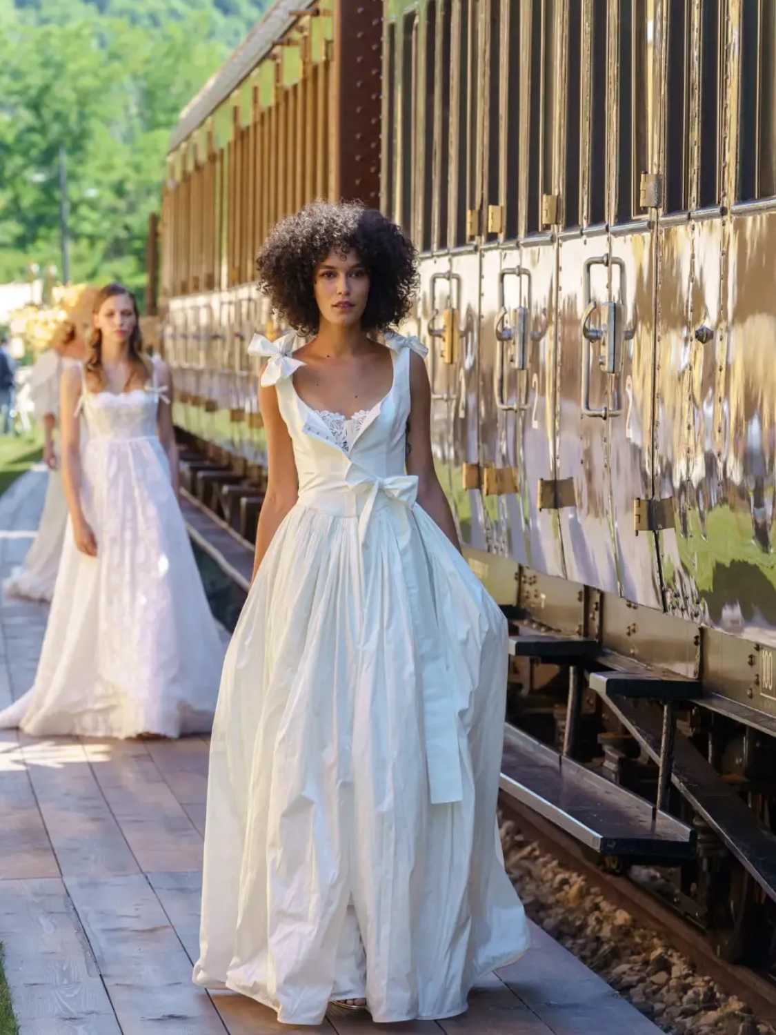 Woman in white dress walks on train platform, another woman in white dress in the background.