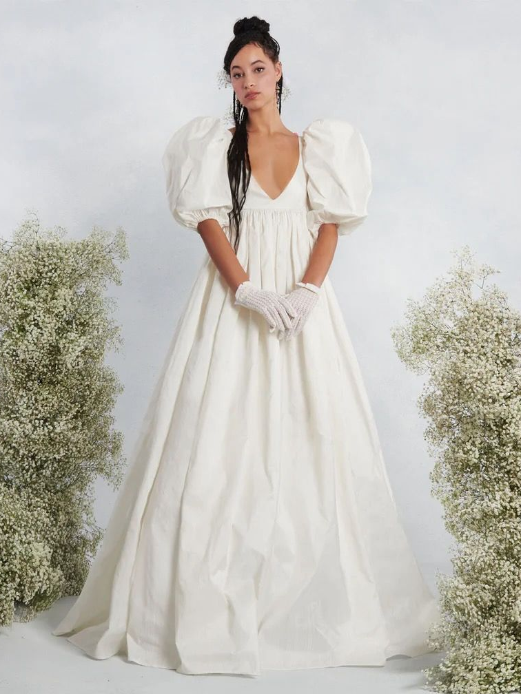 Woman in white dress with puffy sleeves and gloves, stands before baby's breath floral arch.