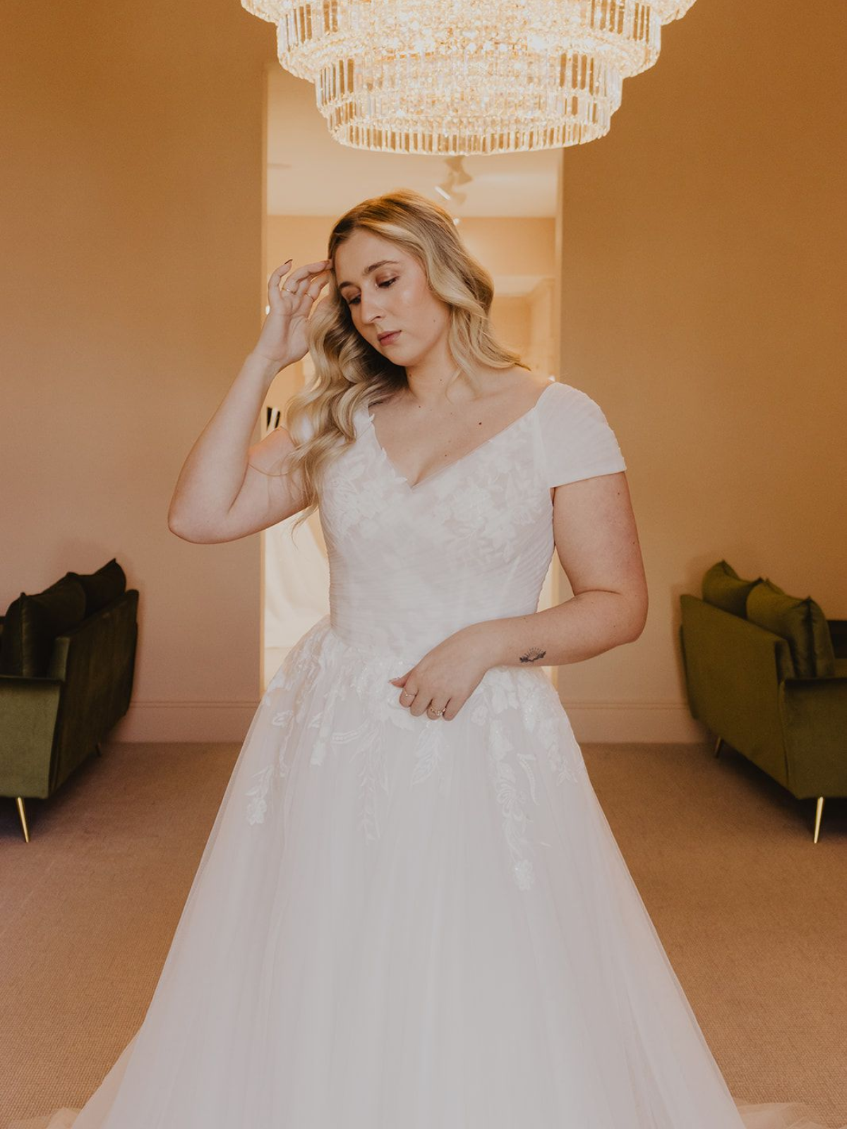 Woman in a white wedding dress stands under a chandelier, hand on her head, looking down.