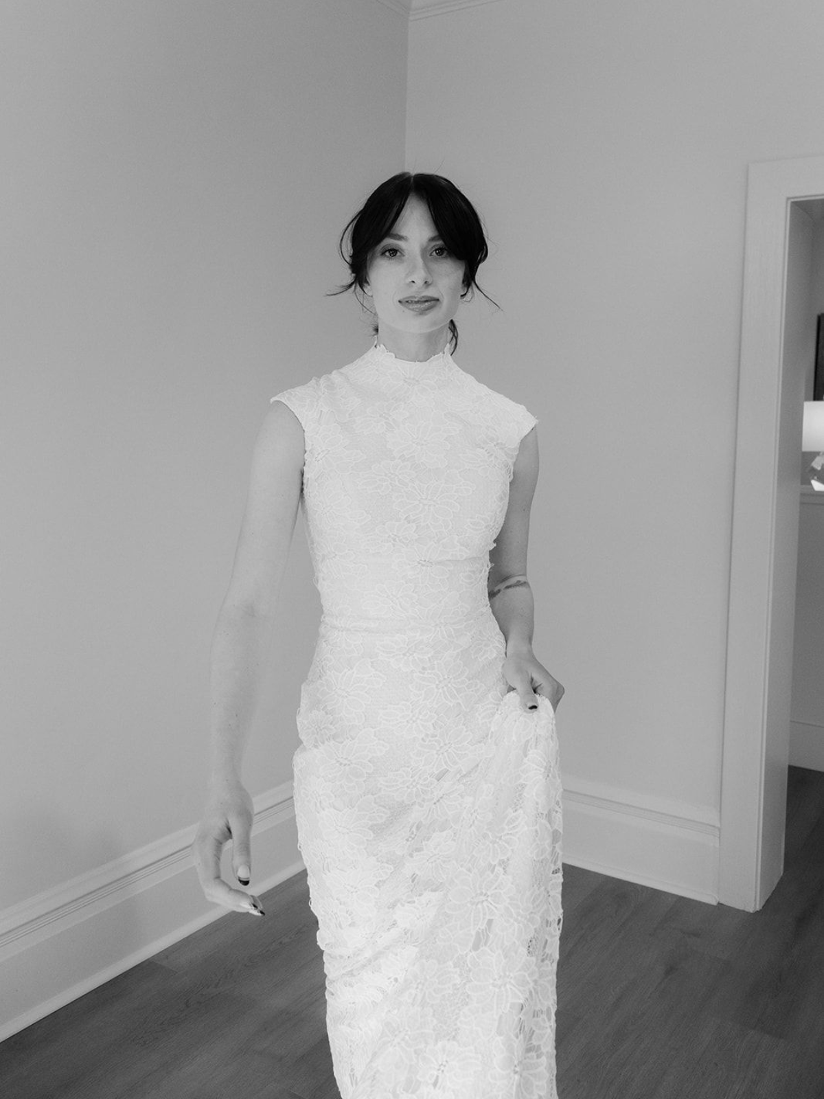 Woman in white lace dress, standing in a room. She has dark hair and a neutral expression.