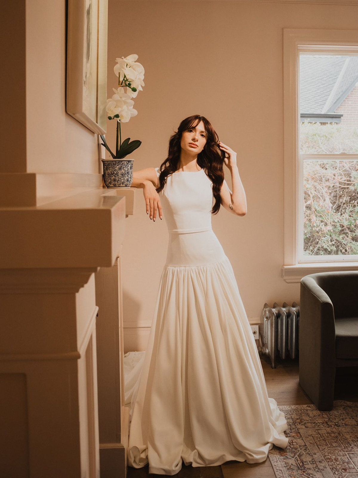 Woman in white gown leans on a mantel. Room with window and potted flowers.