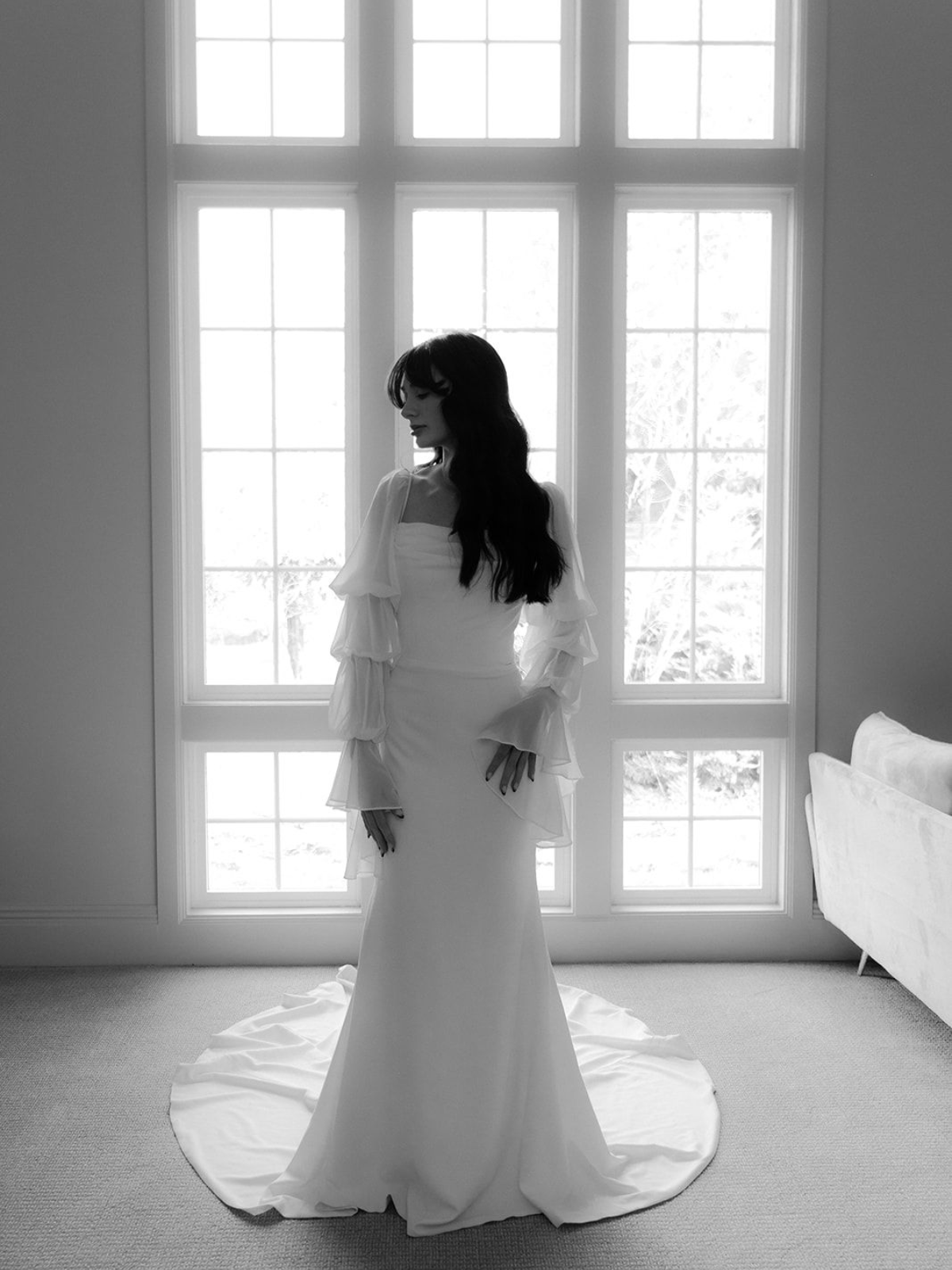 Bride in wedding dress, standing before a large window, with long train trailing on floor.