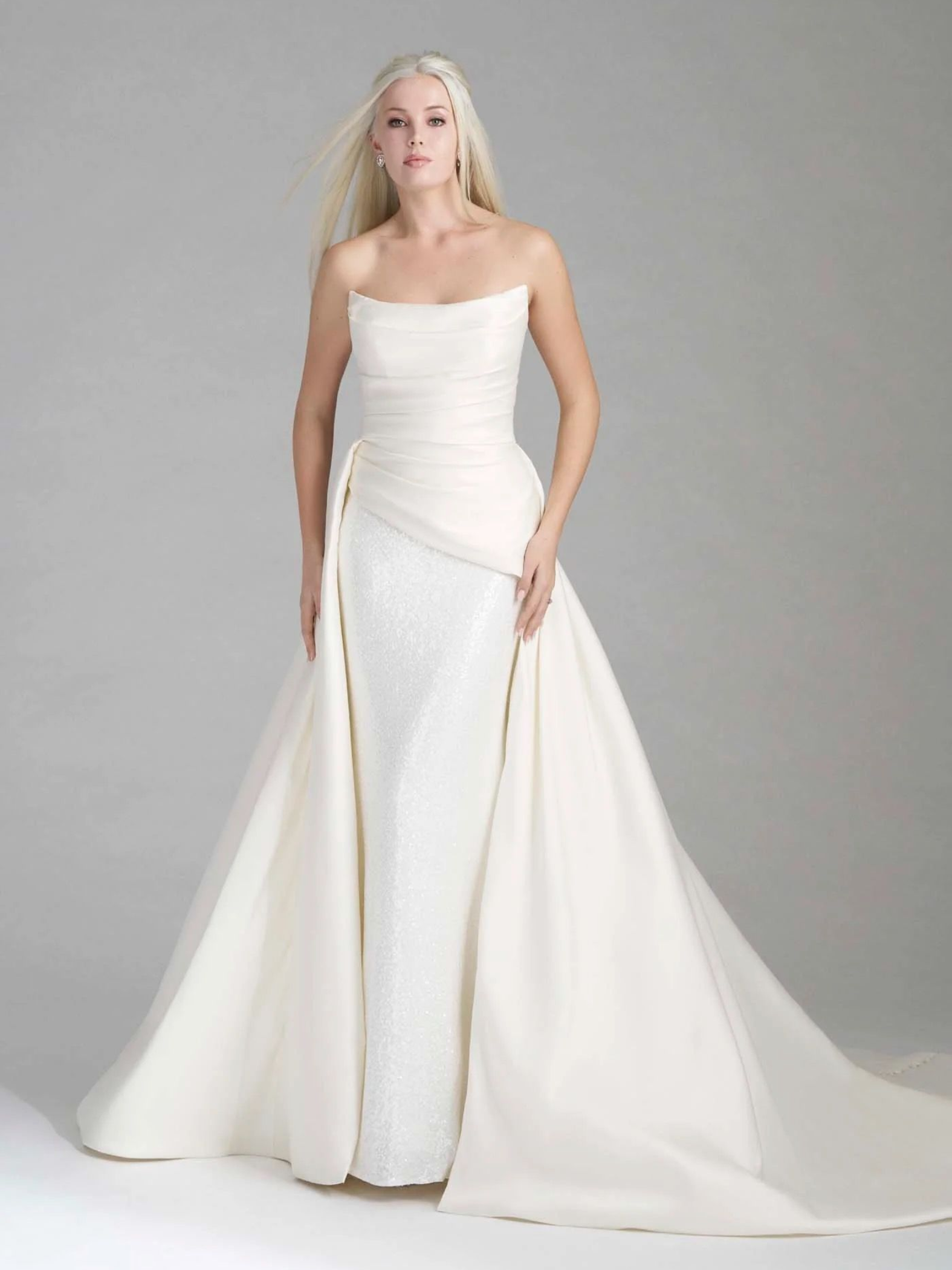 Woman in a strapless white wedding dress with long train, standing against a gray backdrop.