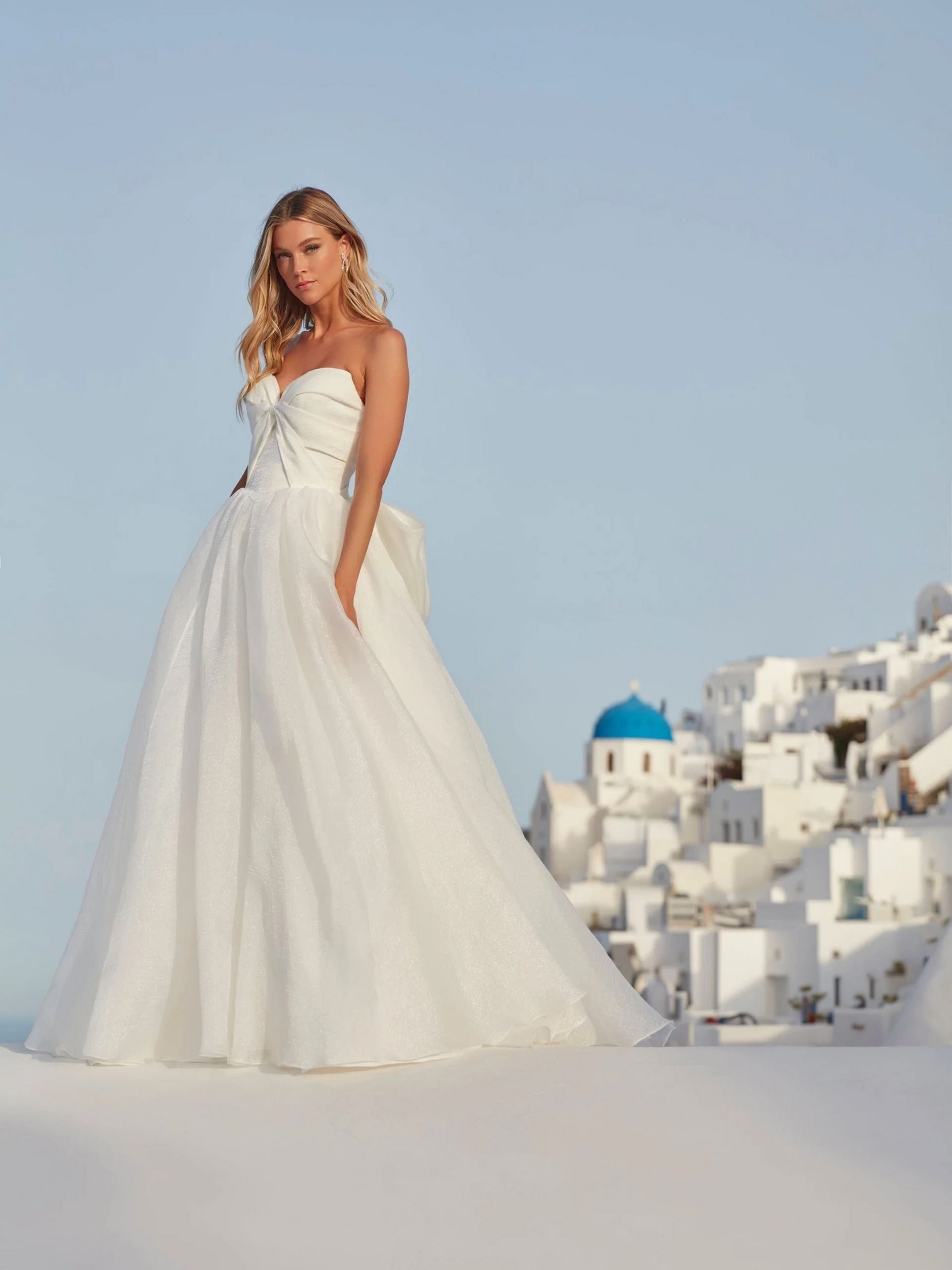 Woman in a white wedding dress posing on a white surface with a background of white buildings and a blue sky.