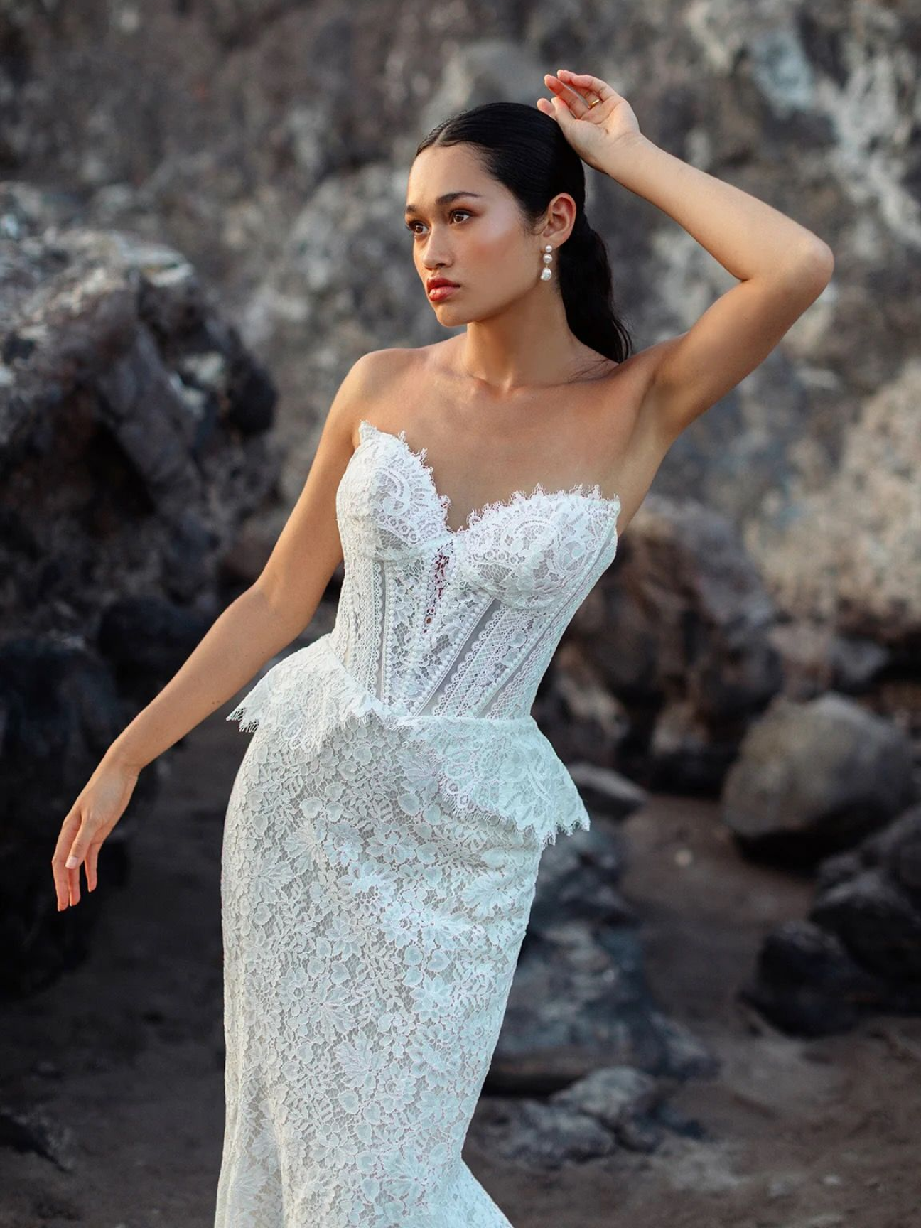 Woman in white lace strapless gown poses outdoors with hand in hair, looking away.