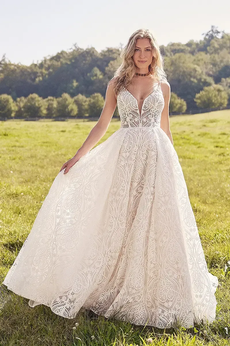 Woman in a white wedding dress stands in a field, holding the skirt of the dress; sunlight shines.