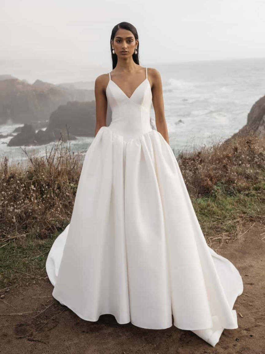 Woman in white satin wedding dress standing near ocean cliffs.