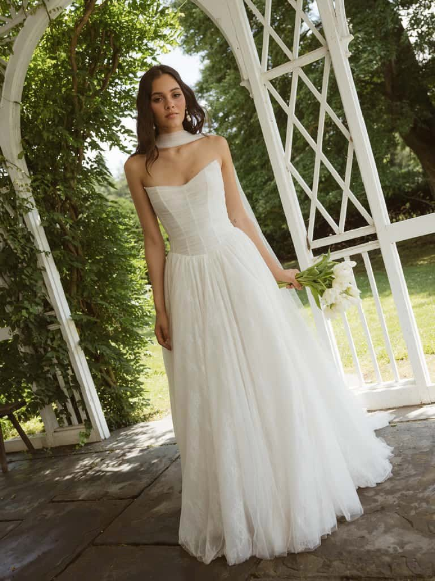 Woman in strapless white dress, holding flowers, standing under white trellis.