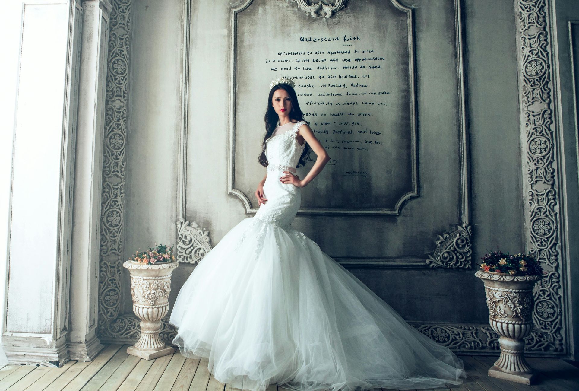 Woman in a white mermaid wedding dress stands in a decorative room with a long train, arm on hip.