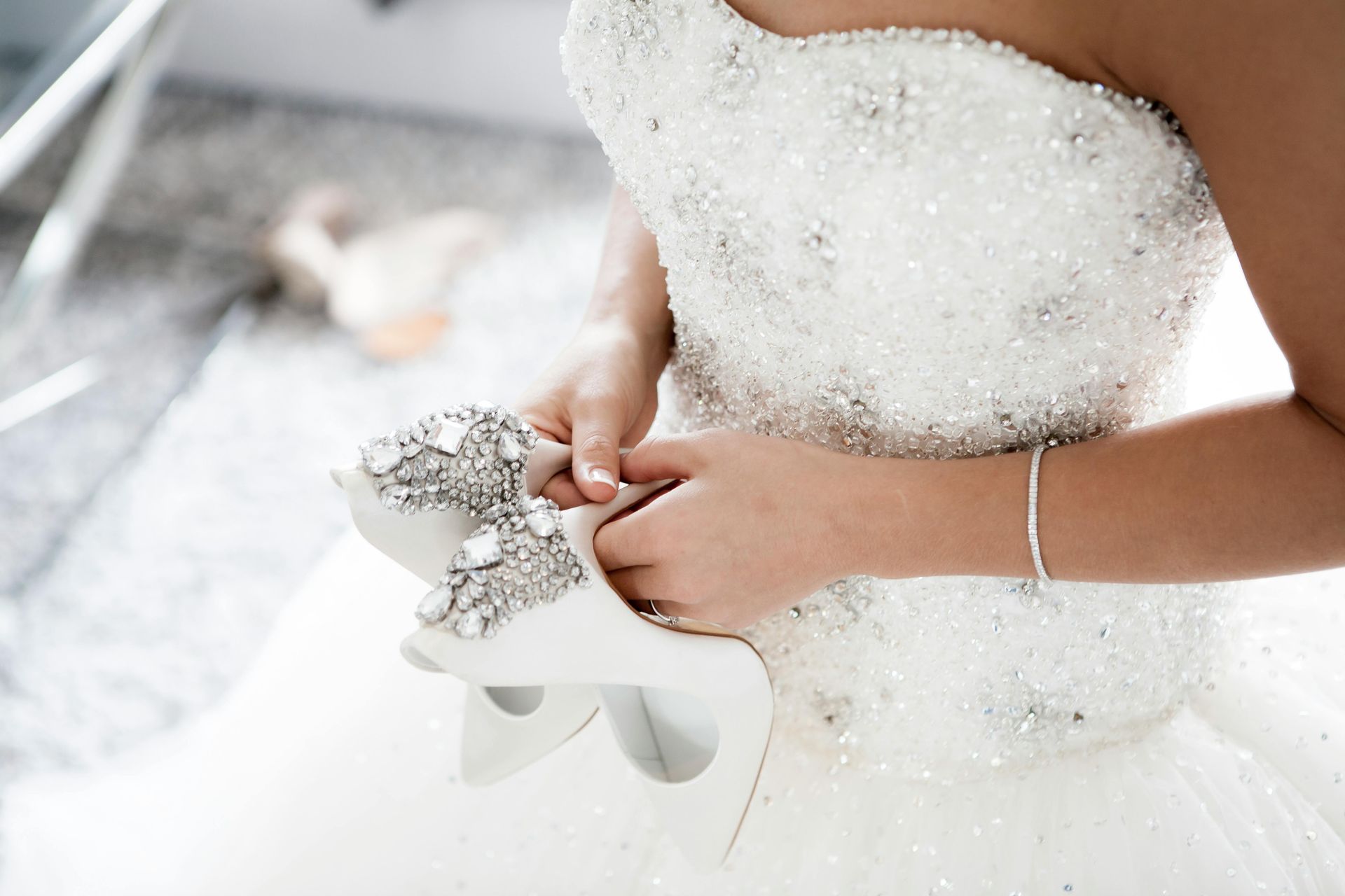 Bride holding white embellished wedding shoes, wearing a beaded gown.