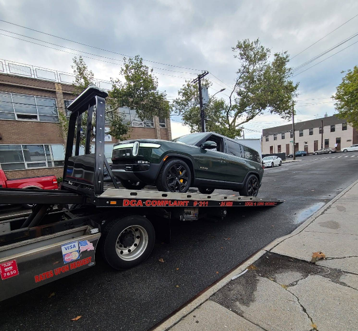 Green Rivian SUV being towed on a flatbed truck on a city street.