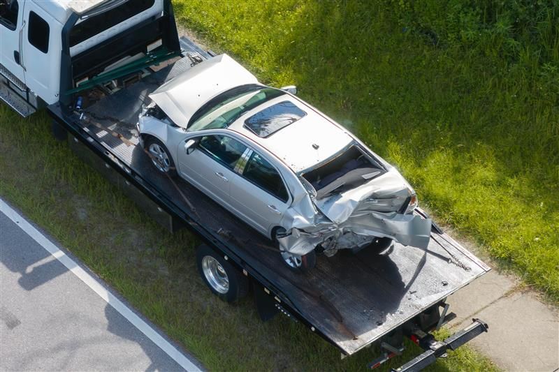 Silver car heavily damaged on tow truck bed, next to grass and road.