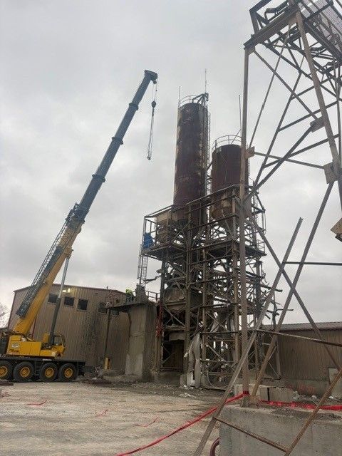 Construction crane lifting steel framework at an industrial site with tall cylindrical towers and scaffolding