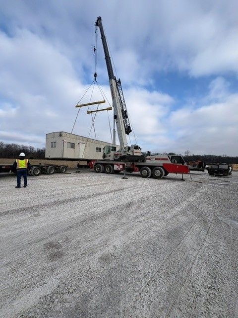 Crane lifting a modular building onto a trailer in a gravel lot under cloudy skies