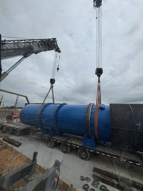 Crane lifting a large blue industrial pipe section onto a flatbed truck at a work site.