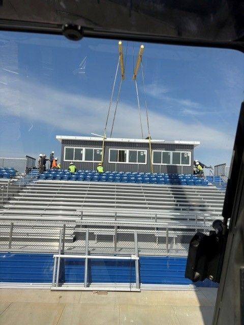 Empty bleachers in a blue stadium with a small press box and workers on the front row