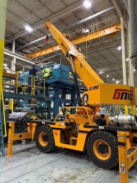 Yellow mobile crane lifting a blue industrial machine inside a warehouse