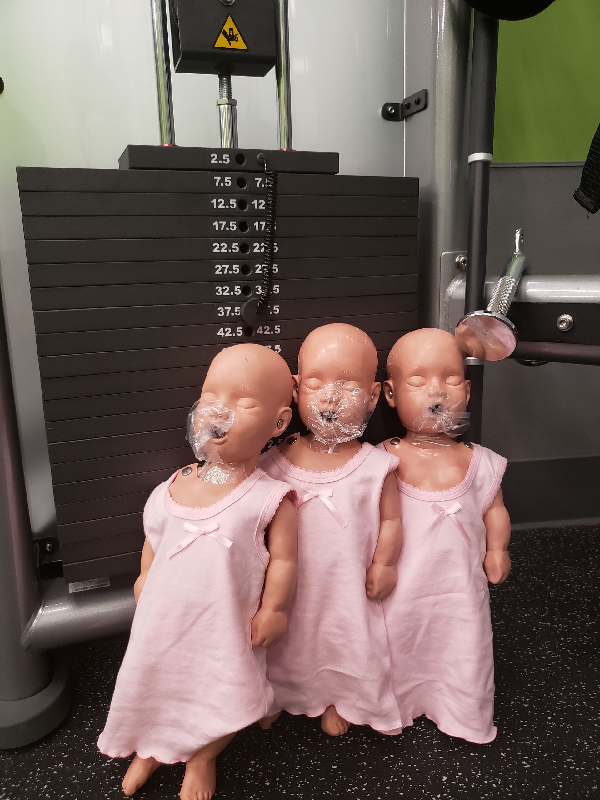 Three dolls in pink dresses standing in front of a gym machine.