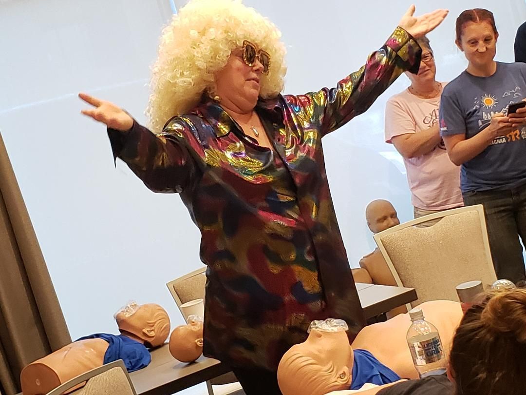 Person in a colorful outfit and blonde wig dancing with arms raised at a gathering, with seated people nearby