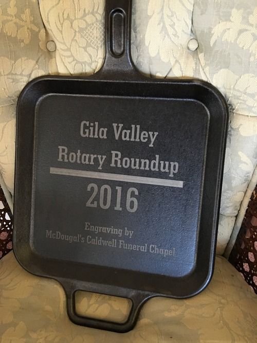 Black cast iron skillet plaque reading “Gold Valley Rotary Roundup 2016” on patterned fabric.