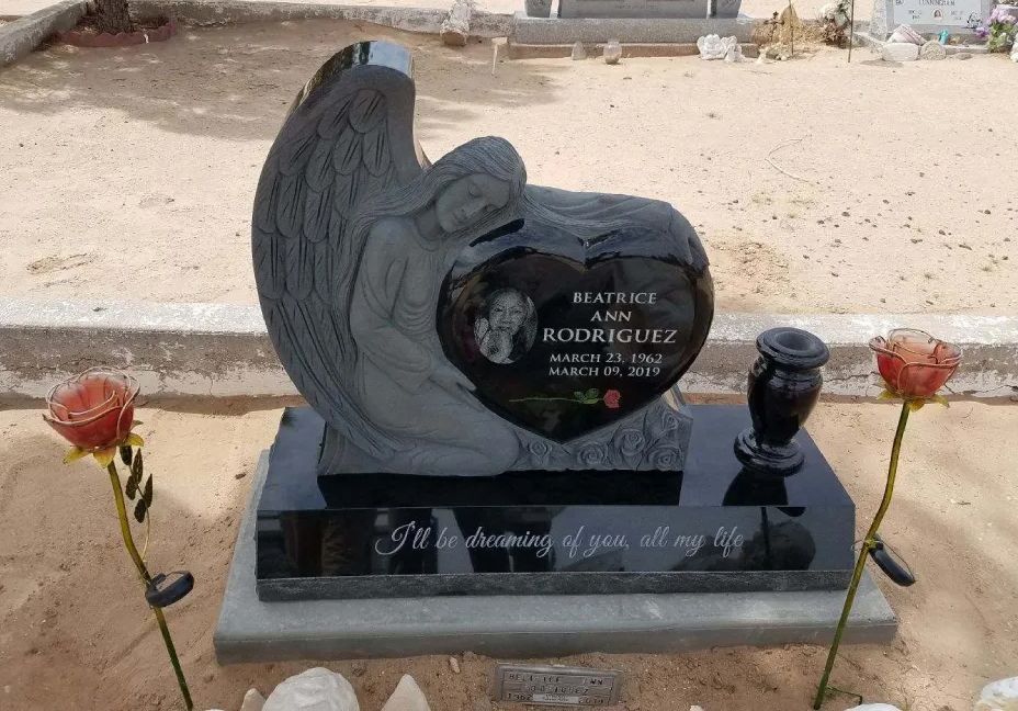Black heart-shaped gravestone with angel wing and red roses in a sandy cemetery.