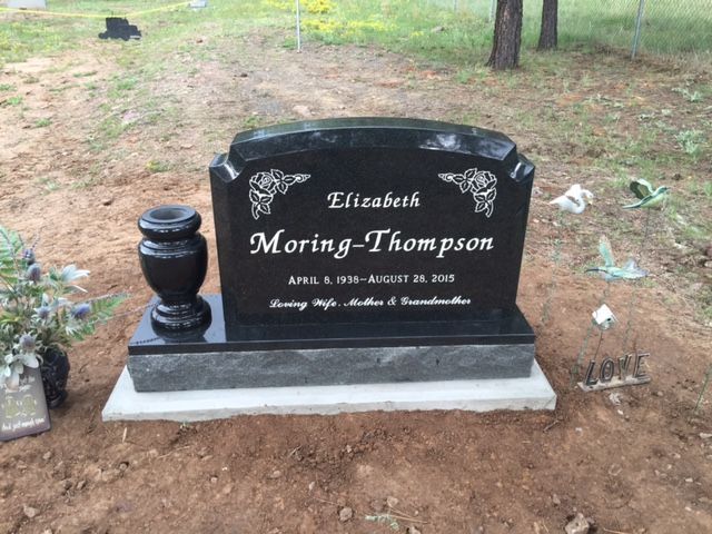 Black gravestone with white lettering on a dirt plot, surrounded by grass and trees.