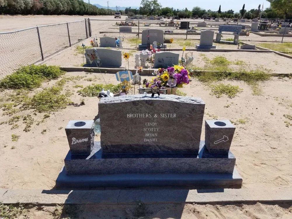 Gravestone with flowers in a sandy cemetery, with other markers in the background.