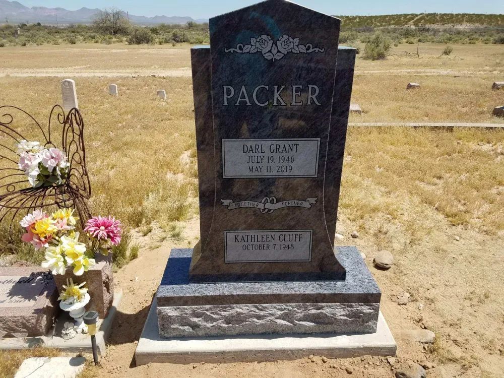 Black granite gravestone in a dry cemetery with flowers beside it under a clear sky