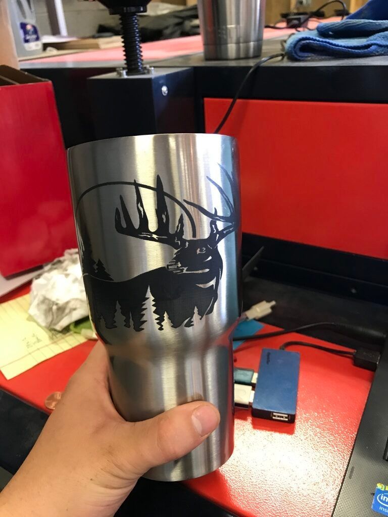 Hand holding a stainless steel tumbler with a black forest and mountain silhouette design.
