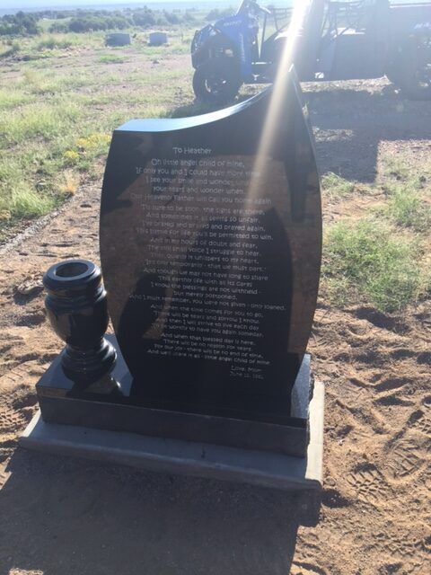 Black memorial headstone with engraved text and a small urn, standing in a sunny graveyard.