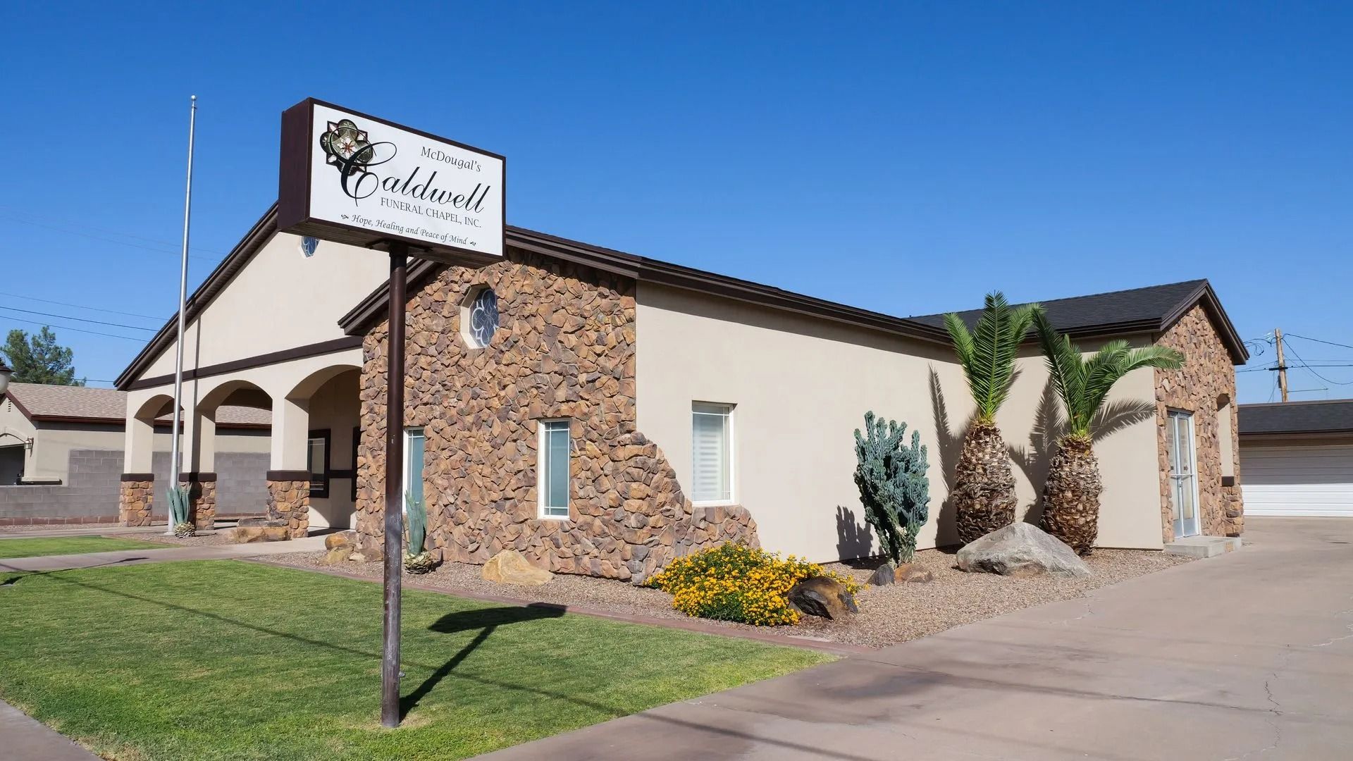 Single-story stucco building with stone facade and sign, front lawn and sidewalk under a clear blue sky