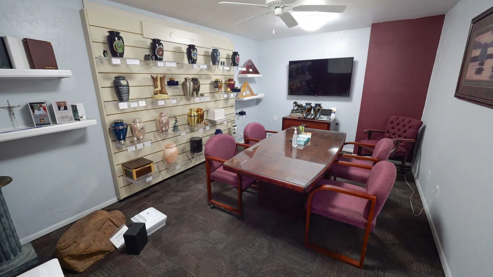 Conference room with long table, red chairs, wall shelves, and a large TV on the far wall.