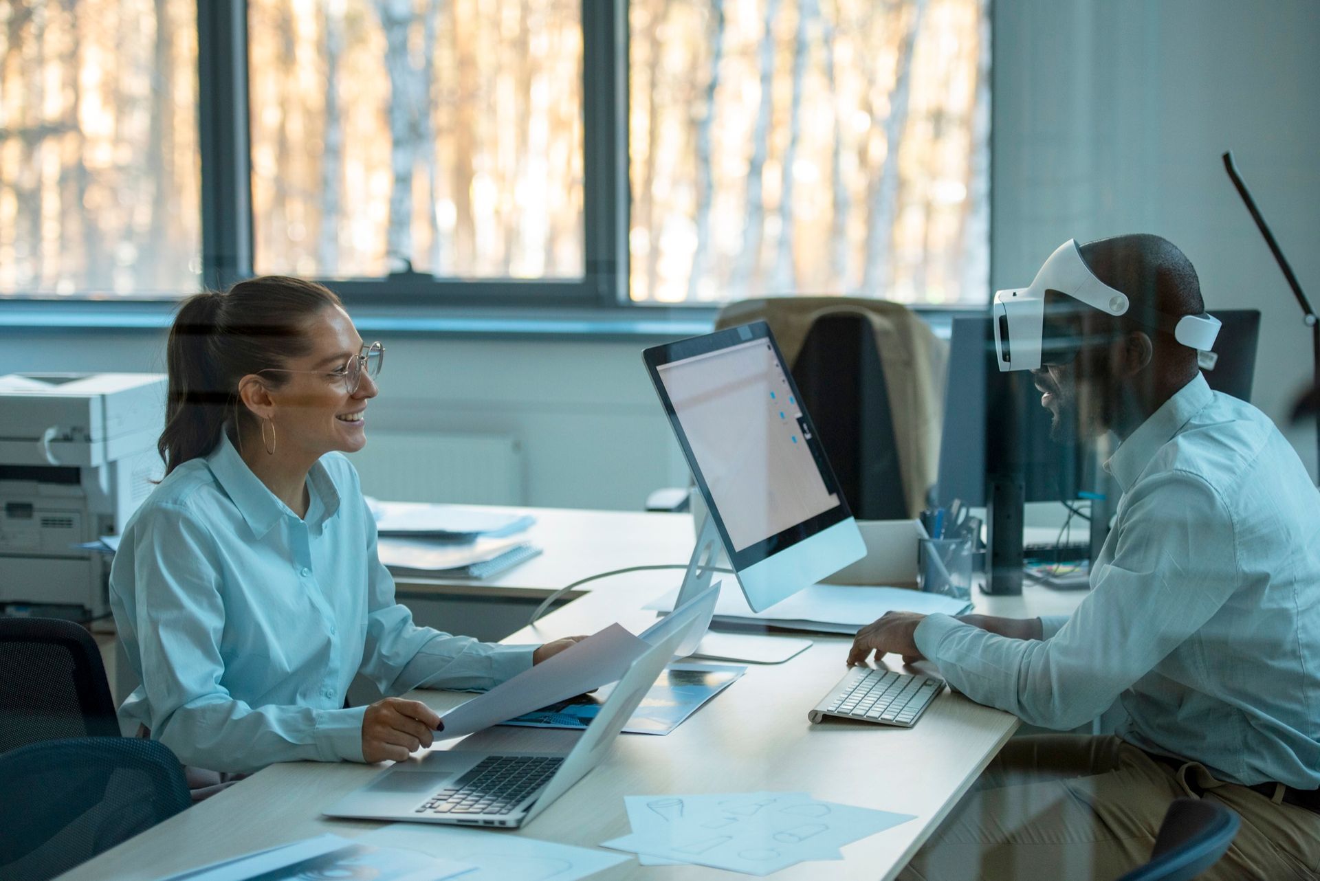 Woman smiling, reviewing documents with a person wearing VR headset, typing at a computer in an office setting.