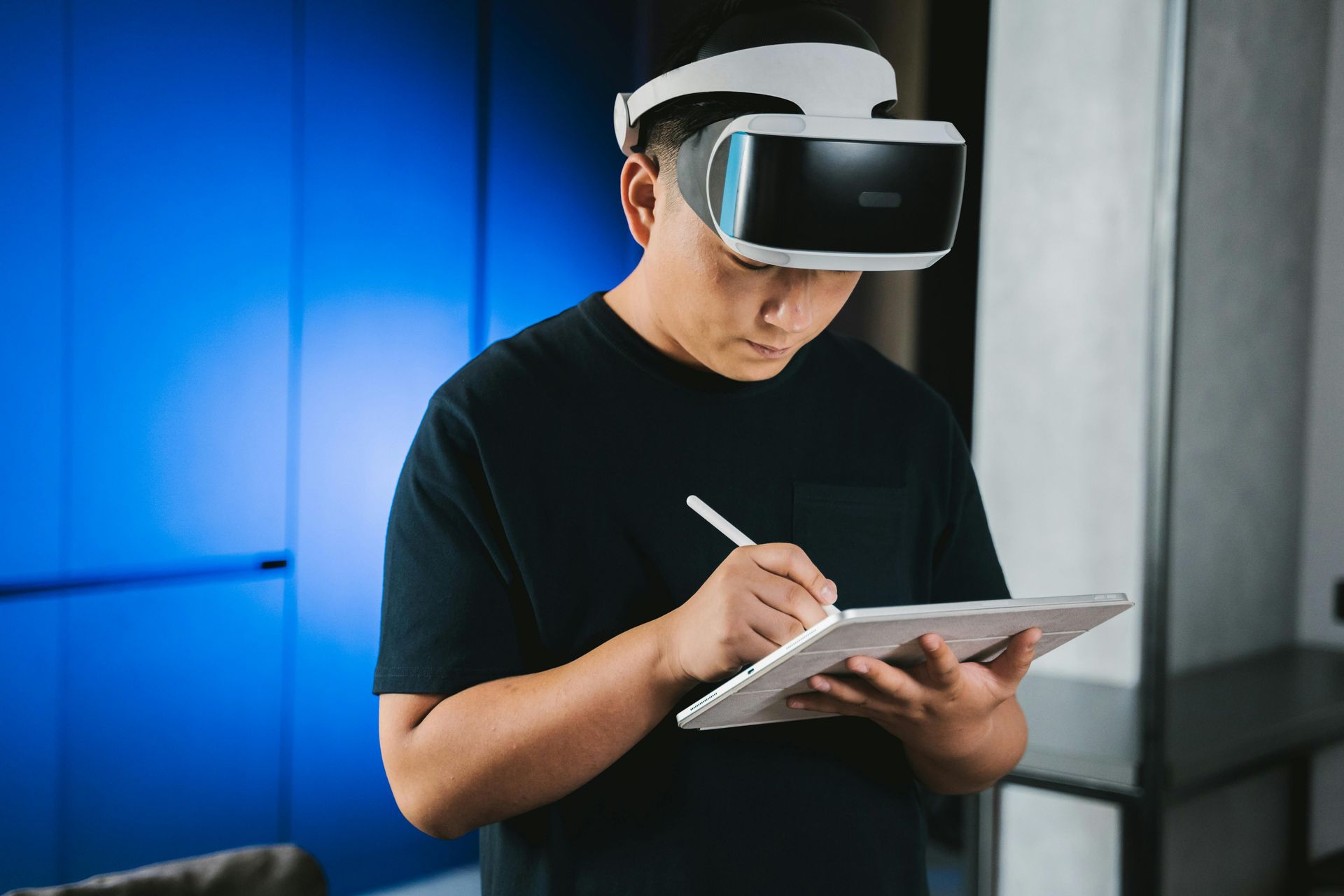 Person wearing VR headset, using stylus on a tablet. Blue wall background.